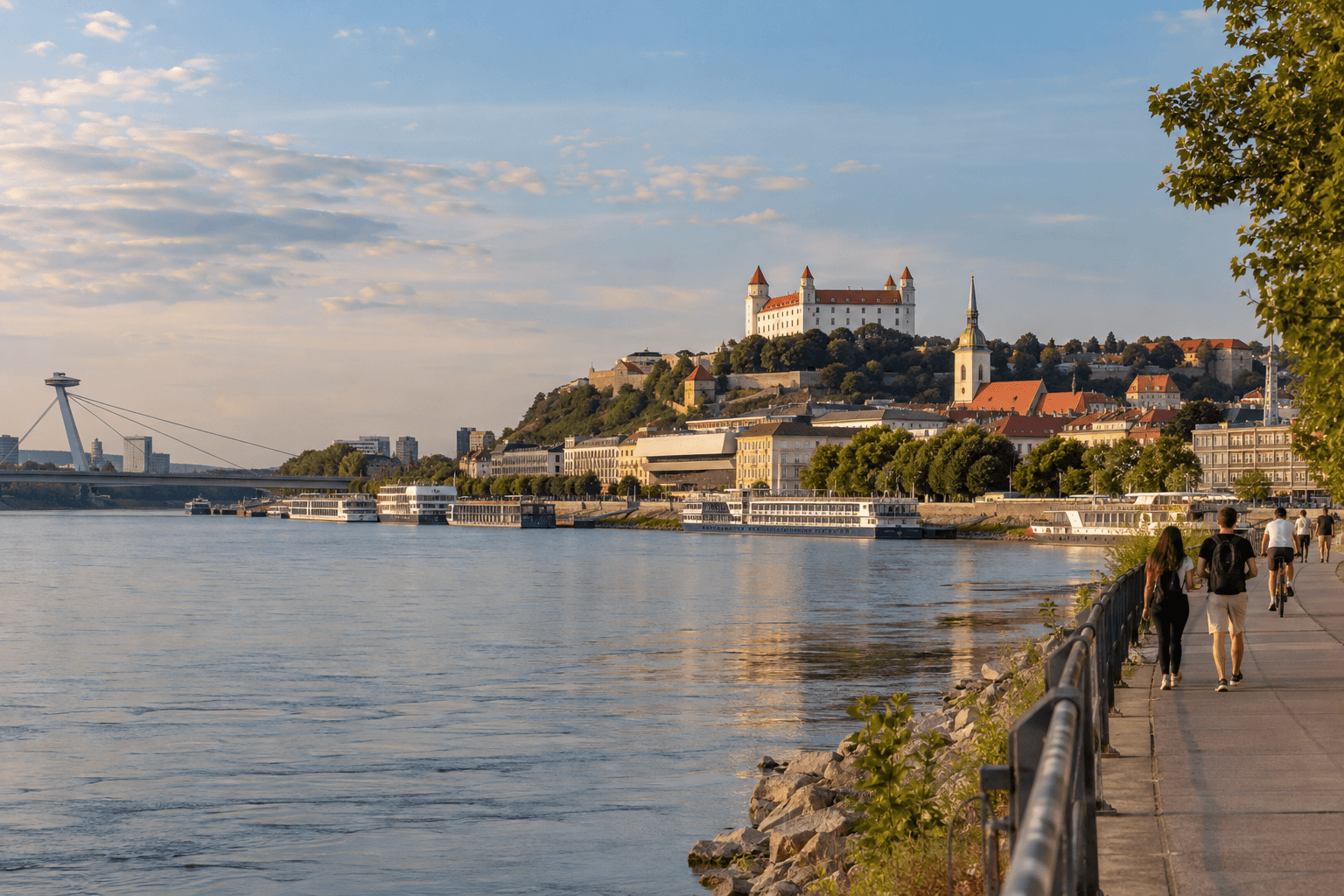 Photorealistic late-afternoon view of Bratislava Castle above the Danube with the Old Town, promenade walkers and cyclists